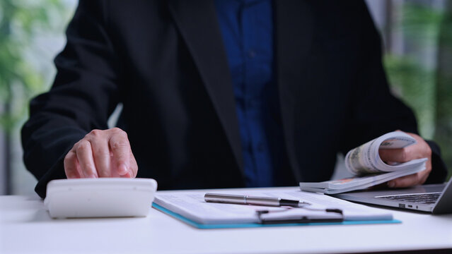 Businessman wearing dark suit counts paper currency near calculator inside office desk workspace, calculating financial budget, managing business tax document, planning investment strategy.