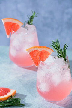Refreshing pink grapefruit mocktail served over ice and garnished with a fresh grapefruit wedge and rosemary sprig. Two elegant glasses of sparkling citrus beverage on a blue textured background.