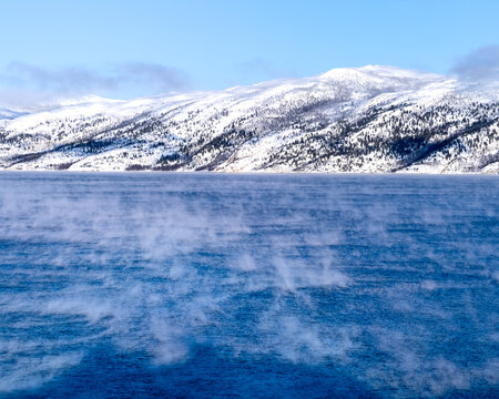 Sea smoke rising from a fjord below a snow covered mountain ridge