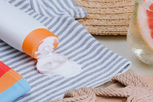 Sunscreen spilled on a towel with stripes. A drink with a slice of grapefruit and herbs sits nearby. Sandals are visible on the side, showing a relaxed beach scene. top view