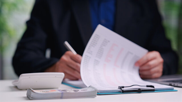 Male accountant analyzes financial document holding pen near calculator stack cash indoor desk workspace, calculating business budget, managing corporate tax expenses, planning investment strategy.