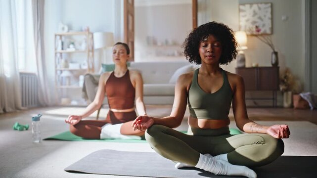 Two women meditating lotus pose home. Multiethnic friends practicing yoga.