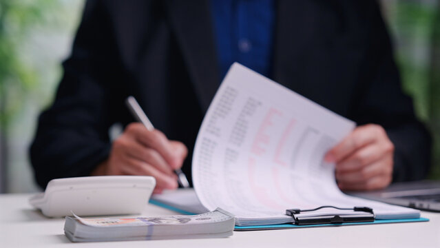 Male accountant reviews financial document, signs clipboard paper, calculates tax budget, manages cash stack, working alone inside office desk, analyzing business investment profit.
