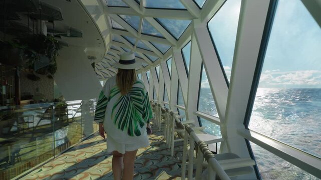 Woman in tropical print shirt and straw hat walks through modern glass-enclosed observation deck on cruise liner in Caribbean Sea. Sunlight sparkles on ocean waves through geometric windows.