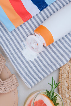 Sunscreen spilled on a towel with stripes. A drink with a slice of grapefruit and herbs sits nearby. Sandals are visible on the side, showing a relaxed beach scene. top view