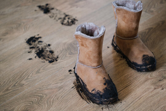 A pair of muddy suede winter boots standing on a wooden floor with a visible trail of wet soil and footprints left behind after coming indoors. Home cleaning, family life, and everyday messes