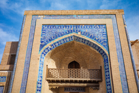 Front view of the magnificent entrance portal of a historic madrasa in Khiva, Uzbekistan, showcasing intricate blue mosaic tilework, traditional brick architecture, and a beautifully crafted wooden