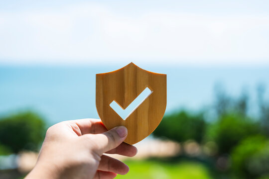 Insurance, security, and guarantee concept. Hand holding wooden shield with check mark against nature background. Ideal for protection, travel insurance, and quality assurance business.