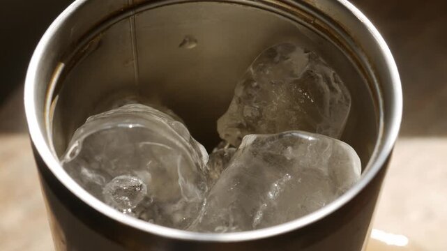 Close-up top angle view into stainless steel tumbler cup with melting ice cubes in golden warm light on wooden table in cafe