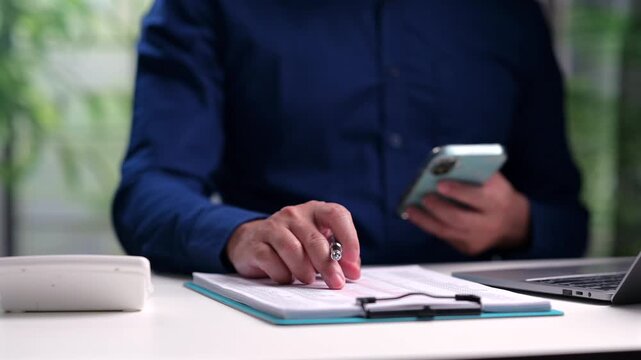 Office worker holding smartphone analyzes paper document, holding pen, sitting desk, reviewing corporate budget reports inside workspace, checking business data, managing company accounts.