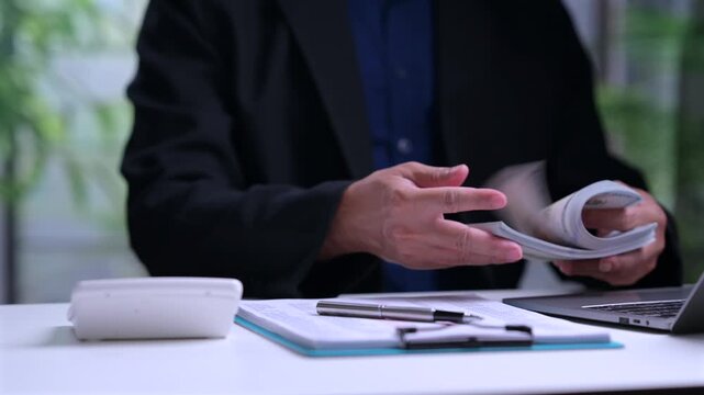 Male entrepreneur wearing suit analyzes financial document near calculator laptop computer, holding cash money stack inside office workspace, planning budget investment profit growth strategy.