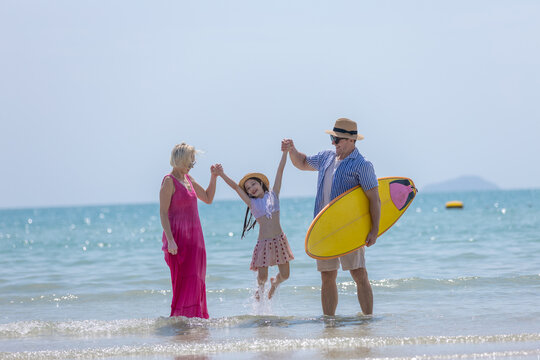 Happy senior family enjoying sunny summer vacation beautiful ocean beach with young child holding hands lifting her with joy carrying yellow surfboard walking shallow water feeling wonderful