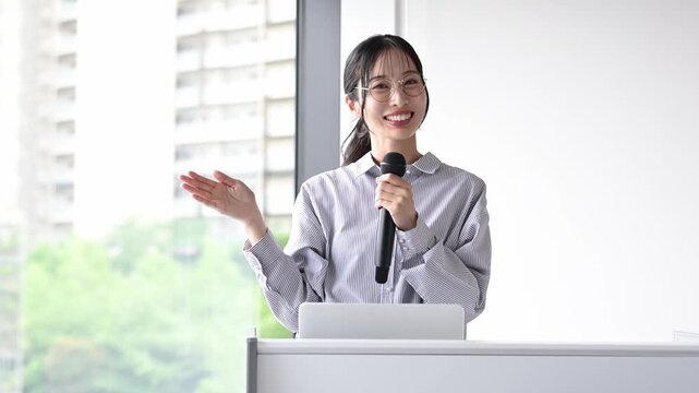 会議室でマイクを持ってプレゼンテーションを行う30代の日本人ビジネスパーソンの女性