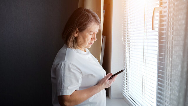 Senior woman standing near a sunlit window with blinds, looking at her smartphone with a serious expression, connecting with technology and the digital world from home