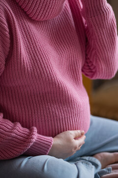 Pregnant woman cradling her belly while seated indoors, wearing a cozy knitted sweater, soft natural light