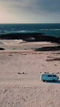 Aerial view showing a retro light blue camper van driving along a deserted sandy beach next to a beautiful turquoise ocean with waves breaking on the rocky coastline, representing freedom