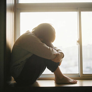 Depressed person sitting on window sill with head down in dark home interior space.