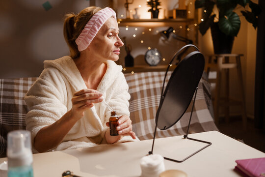 senior woman wearing bathrobe using facial oil or serum sitting in front of mirror, testing face care product cosmetics in evening routine.
