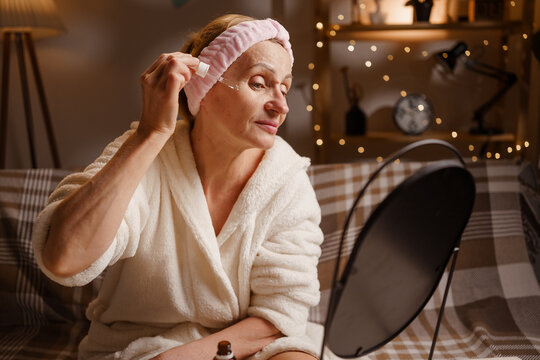 senior woman wearing bathrobe using facial oil or serum sitting in front of mirror, testing face care product cosmetics in evening routine.