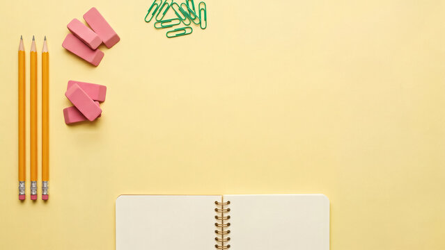 Pencils erasers paper clips and a spiral notebook arranged on a pale yellow surface stationery writing