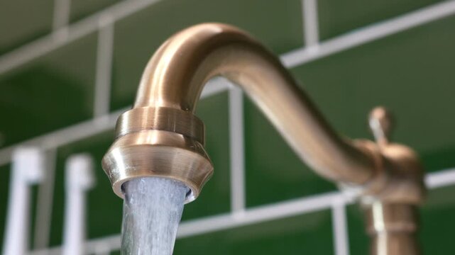 Water flowing from a vintage bronze faucet against a green tiled wall background. Concert - payment of utility bills for water, water consumption in the apartment, clean water from the tap. High quali