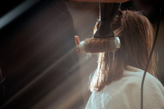 Close Up Of Hair Wrapped Around Round Brush With Dryer At Salon. Back view of a woman with towel on shoulders getting a professional blow dry styling treatment