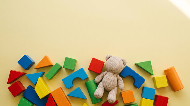 Knitted teddy bear surrounded by vibrant geometric wooden building blocks on soft yellow background with assortment colorful wooden various geometric shapes
