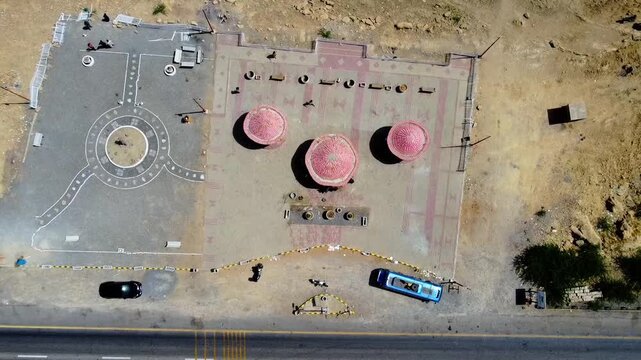Drone video captures vibrant roadside vendor huts, scattered people, and numerous fishing boats at Kund Malir Beach, Balochistan. Bright sunlight, arid terrain, and busy shoreline are visible.