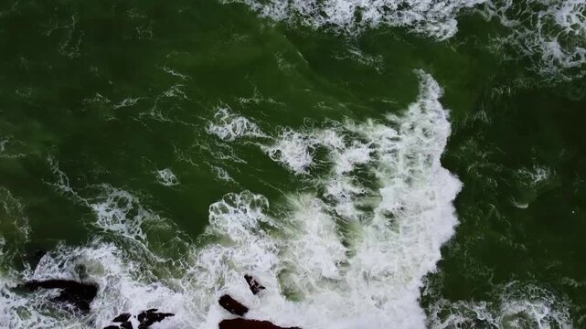 Stunning aerial footage of green ocean waves crashing against rugged brown rocks at Mubarak Beach, Karachi. Dramatic dynamic movement, swirling foam, and textured coastline under daylight.