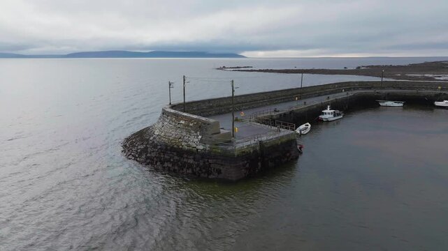 Cinematic aerial shot of a traditional stone pier in Barna, Ireland, featuring small boats tied to the harbor under a moody overcast sky.
