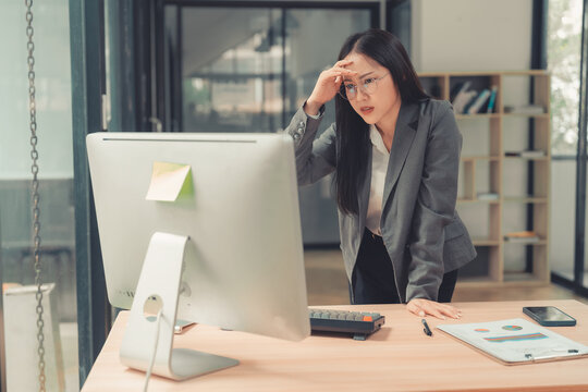 Stressed Asian businesswoman holding hand on forehead looking at computer screen with worried expression.