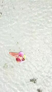 Top down aerial view of a young relaxed woman in a bikini sunbathing on a colorful inflatable mattress, floating on the calm, transparent surface of a shallow turquoise sea during her vacation