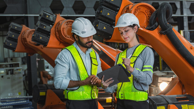 Professional engineers reviewing maintenance documentation together while inspecting industrial robotic equipment in an automated production environment.