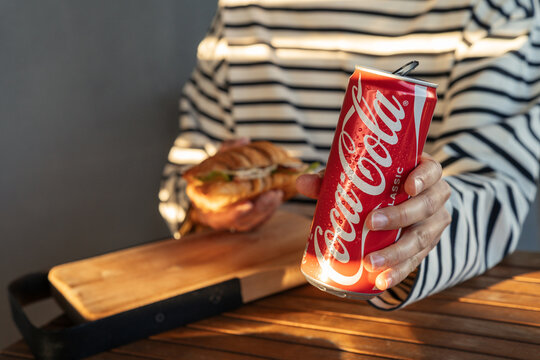Minsk, Belarus - July 30, 2026: Horizontal shot of a person in a striped top holding an opened can of Coca-Cola Classic and a croissant sandwich over a wooden table
