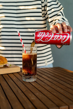 Minsk, Belarus - July 30, 2026: Pouring Coca-Cola from a red can into a tall glass with a paper straw over a wooden table