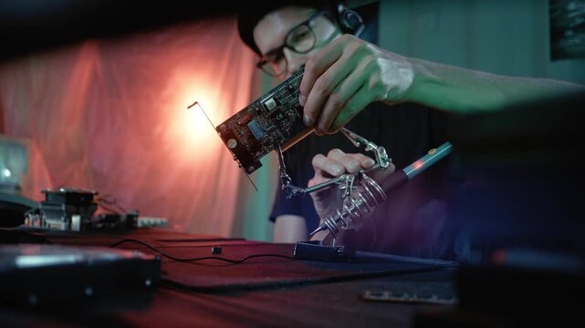 Male hacker wearing a headset soldering a computer video card in a dark room with green and red light