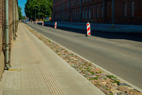 City street under construction with red and white traffic cones on asphalt road. Empty sidewalk next to brick buildings.