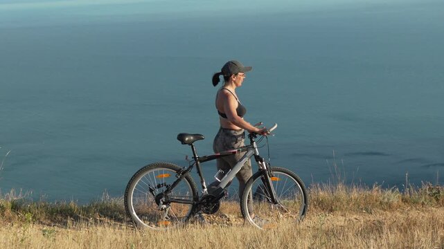 Bicycle travel adventure, a woman standing with mountain bike on a cliff overlooking the ocean during a sunny summer day in nature