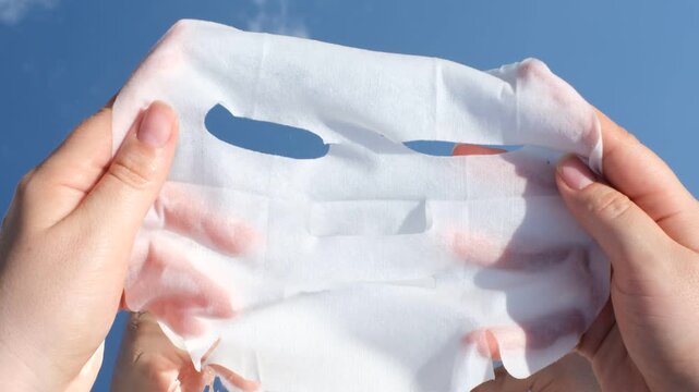 Female hands holding a mask sheet on blue sky and clouds background close-up.
