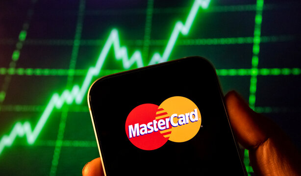 Mastercard logo on phone screen with financial stock market graph background.