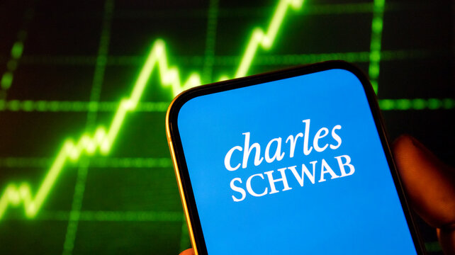 person holding smartphone with charles schwab logo, stock market chart displaying green uptrend in background.