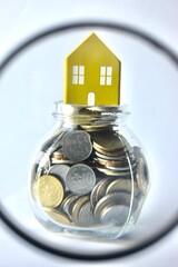 View through a magnifying glass looking at a miniature yellow toy house placed on top of a coin-filled glass jar, representing property investment analysis, real estate evaluation, and housing market. © Akram