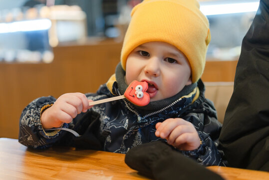 A joyful child eats a lollipop and tastes a sweet treat at a table in a cozy cafe. The curious toddler holds the candy with excited delight from a lowangle view in warm restaurant lighting.