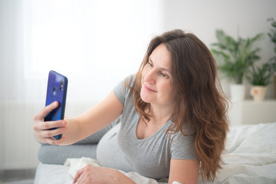 Pregnant woman taking selfie while lying happily in bed. Expecting mother holding blue phone with carefree smile in casual bedroom. Modern mom capturing joyful moment on white sheets near plant.