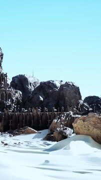People explore large rocks and old structures in a desert landscape. The sun shines down on the white sand and stone formations. The clear blue sky frames the scene above.