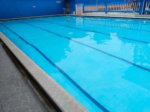 Clean outdoor lap swimming pool with bright blue water and lane lines. Stainless steel pool ladder in foreground, tiled deck and blue walls in background