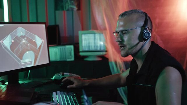 A male computer operator works with a simulated security camera system while communicating through a headset in a dark neon-lit container workspace.