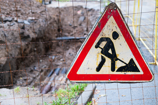 A warning sign shows a construction worker digging at a site in a busy urban area. The scene includes fencing and excavated ground, signaling that work is happening and detours may be needed