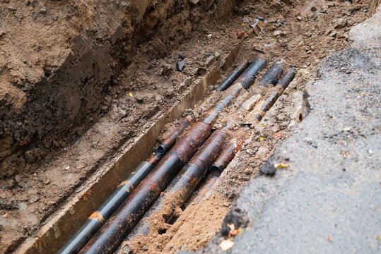 conducting repairs in a trench that exposes several underground pipes. The site shows dirt and debris from the digging process, indicating ongoing maintenance activities