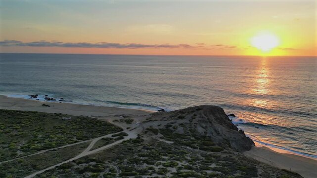 Drone lowers and pushes in toward the rocky point of Secret Rocks Beach below Todos Santos, watching waves wash ashore beneath a glowing Pacific sunset.
